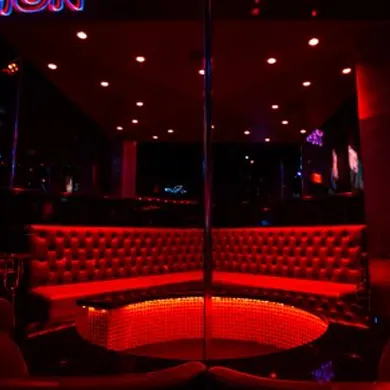 A gentleman's night out setting in a dimly lit club, featuring a central stage with a pole, surrounded by glowing red seating. The mood is intimate and exclusive.
