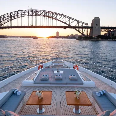 An aerial view from the deck of a yacht shows the Sydney Harbour Bridge at sunset. The wooden deck has lounge chairs and tables. The water is calm and reflects the orange and yellow sky.