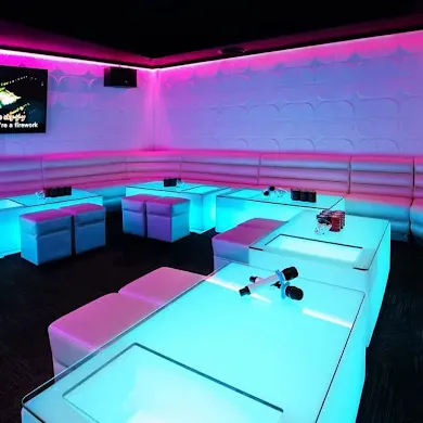 A vibrant karaoke room for a lively karaoke night, featuring white padded seating and glowing tables illuminated with bright pink and blue LED lights. The walls have a textured, wavy pattern, and a large screen for lyrics is visible in the corner.