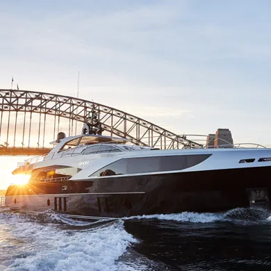 A luxury yacht cruises past the Sydney Harbour Bridge at sunset, with the sun's rays reflecting off the water.